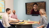 A photograph of a meeting space. At the front of the room, against the wall, there is a TV screen showing an active Google Meet call. A Logitech MeetUp 2 is mounted below the screen. In front of the screen, there is a semicircular table with a Logitech Tap placed on top and three people sitting around the table in chairs.