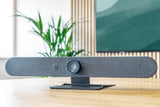 An image of the Logitech Rally Bar Mini on a table in a meeting room. Behind the Rally Bar Mini, there is a TV screen mounted on the wall.