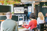 An image of an open meeting area where four participants are facing towards a TV screen showing an active Google Meet call. The screen is mounted on a cart along with the Logitech Rally Bar and the Logitech Tap.