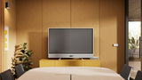 An image of a meeting room with Logitech Rally Board 65 mounted on a credenza using table legs at the front of the room. The built-in camera is situated below the display and a large table surrounded by chairs is in the foreground.