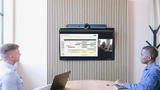 A photograph of a meeting space with a Logitech Rally Bar mounted above a TV screen on the wall. There are two people sitting at a table, facing the TV screen at the front of the room.